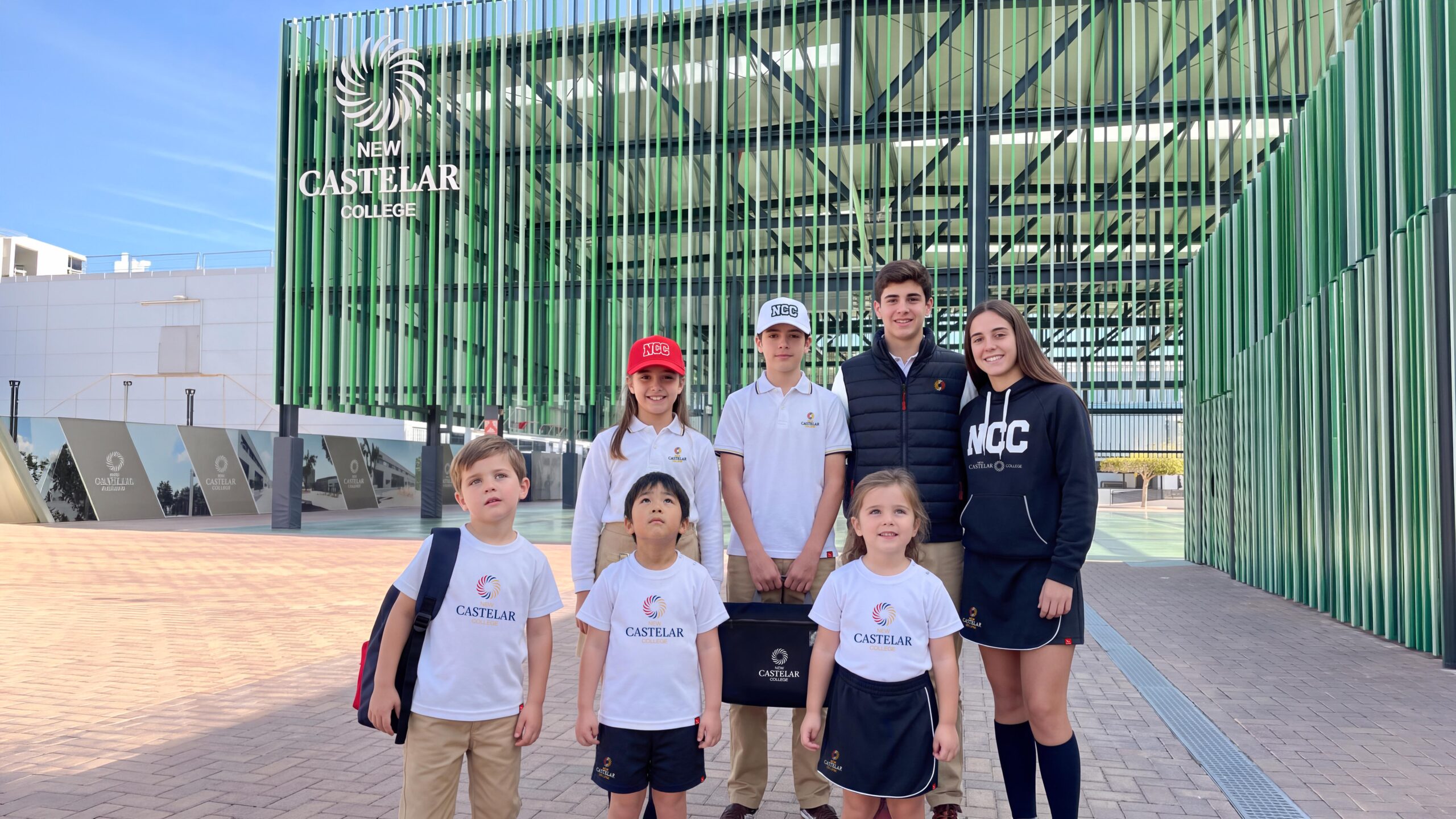 Grupo de estudiantes de varias edades posando en la entrada de New Castelar College vistiendo chalecos, sudaderas, polos y gorras personalizadas.